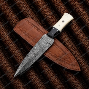 May include: A fixed-blade knife featuring a pointed Damascus steel blade with a wavy pattern, a black bolster, and a cream-colored handle with two metal pins. The knife rests on a brown leather sheath embossed with a decorative leaf pattern.