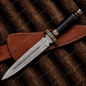 May include: A Damascus steel dagger with a black handle and ornate gold-colored accents. The dagger rests in a brown leather sheath, set against a textured brown background. The blade has a distinctive pattern.