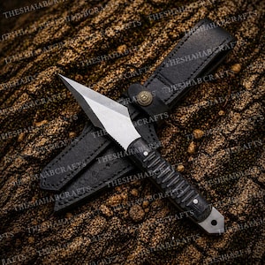 Könnte beinhalten: Ein kleines Messer mit silberner Klinge, strukturiertem schwarzem Griff und schwarzer Lederscheide. Das Messer hat eine spitze Spitze und eine gezahnte Kante in der Nähe des Griffs. Die Scheide hat eine Gürtelschlaufe und einen dekorativen Knopfverschluss. Das Messer und die Scheide werden auf einer strukturierten, braunen Oberfläche präsentiert.