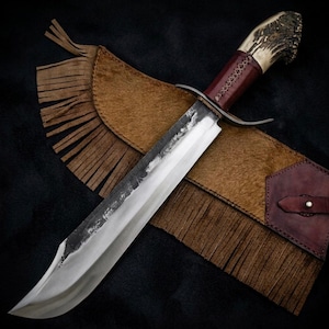 Peut inclure: Un grand couteau de chasse fait à la main avec une lame en acier poli et un manche en corne et cuir rouge foncé. Le couteau repose sur un étui en cuir marron avec des franges.