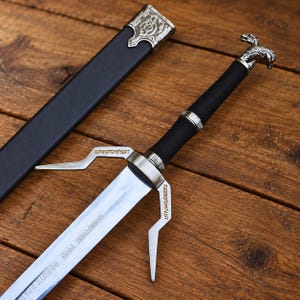 May include: A silver-toned steel sword with a black wrapped handle and a wolf-shaped pommel. The crossguard features angular metal extensions with engraved text. A matching black scabbard with silver-toned metal fittings lies beside the blade.