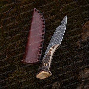 Puede incluir: Un cuchillo de hoja fija con un patrón hexagonal texturizado en la hoja de metal y un mango de asta natural, acompañado de una funda de cuero marrón con bordes cosidos oscuros, sobre una superficie de musgo oscuro.