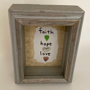 May include: A gray wooden shadow box with a distressed finish. Inside, a piece of paper displays the words "faith, hope, love" with heart-shaped and teardrop-shaped accents in green, white, and brown. The box is a decorative item.