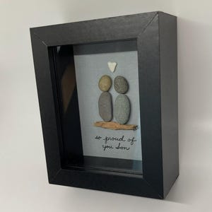 May include: A framed pebble art piece with two figures holding hands, a small white heart above them, and the text "so proud of you son" below. The frame is black and the background is a light gray fabric.