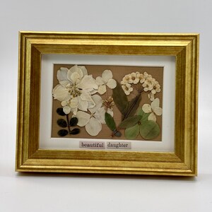 May include: A framed floral art piece featuring pressed white flowers and green leaves on a tan background. The gold frame surrounds a white mat and a small label that reads "beautiful daughter."