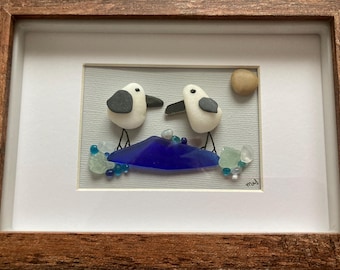 May include: Framed artwork featuring two stylized white seagulls with black accents, standing on a blue glass wave. The artwork includes a small, round, tan stone representing the sun, and is framed in a rustic brown wooden frame.
