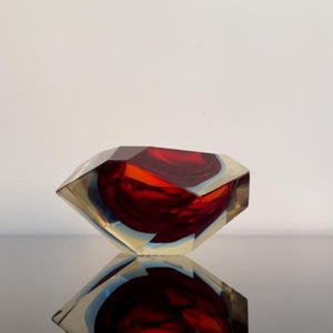 Puede incluir: Un cuenco de vidrio geométrico facetado con un interior rojo intenso y un exterior transparente. La forma del cuenco se refleja en la superficie negra brillante de abajo. El vidrio tiene una apariencia en capas, con toques de azul y amarillo.