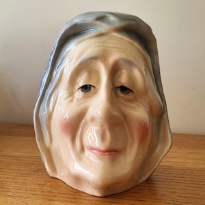 May include: A ceramic figurine of an elderly woman's face. The face is painted with realistic details, including blue eyes, rosy cheeks, and gray hair. The figurine has a slightly smiling expression and is set against a neutral background.