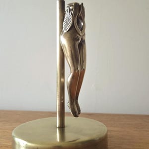 Könnte beinhalten: Eine messingfarbene Metallskulptur, die eine stilisierte menschliche Unterkörperform zeigt, welche an einem vertikalen Stab auf einem runden Sockel befestigt ist. Die Figur weist detaillierte Bein- und Hüftkonturen sowie eine blattartige Textur am oberen Ende auf.