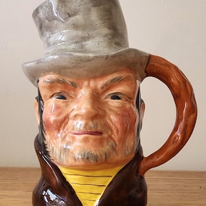 Könnte beinhalten: Ein Keramikkrug in Form eines Männerkopfes mit Zylinder. Der Krug hat einen braunen Henkel und ist mit Gesichtsdetails, einem gelben Hemd und einem braunen Mantel bemalt. Der Hut ist grau.
