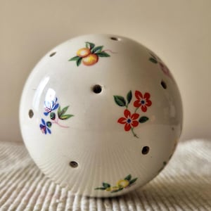 May include: A spherical white ceramic object featuring multiple small circular holes distributed across its surface. The exterior is decorated with colourful floral and fruit motifs, including red flowers, blue blossoms, and small orange fruits.
