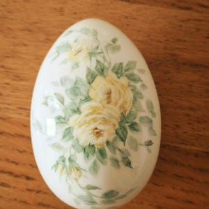 May include: A smooth, white, egg-shaped porcelain object featuring a delicate, hand-painted floral design. The pattern consists of soft yellow roses with muted green leaves and stems, arranged in a cluster that covers the center of the surface.