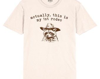 May include: A beige t-shirt with a brown raccoon wearing a cowboy hat and the text "actually, this is my 1st rodeo".