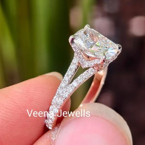 Puede incluir: Un anillo de tono plateado con una gran piedra central rectangular de corte radiante y una banda de vástago dividido incrustada con pequeñas piedras redondas en pavé. El texto Veena Jewells es visible en el lateral del engaste.