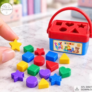 Puede incluir: Un cubo clasificador de formas de plástico rojo y azul con asa y tapa con recortes para un cuadrado, corazón, estrella, círculo y triángulo. Una colección de pequeñas formas de plástico de colores se encuentra junto al cubo.