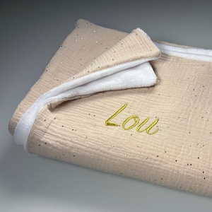 Peut inclure: Une couverture de bébé beige avec des paillettes dorées et une bordure en peluche blanche. Le nom "Lou" est brodé en fil d'or. La couverture est pliée, révélant la texture douce du tissu.