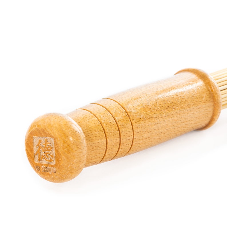 K&ouml;nnte beinhalten: Ein Holzschneebesen mit einem braunen Griff und einem nat&uuml;rlichen Bambus-Schneebesenkopf. Der Griff hat ein quadratisches Logo mit dem Text "yu designs" und einem japanischen Schriftzeichen.
