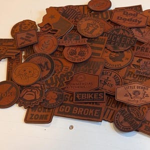May include: A collection of brown leather patches with various designs and text, including "Dad Daddy", "Little Debbie", "Go Broke", "Rump 2024", "Holy Bible", "Son", "Jesus", "EBikes", "Commercial Kitchens", "Ride", "No Worries", "Try That", "Work Zone", "Premier Custom Leather", "Duckers", "Prestige Construction", "Happy Tail", "Stand Out", "62", "Mowrks", "Romp", "Zzy", "Oke", "With Us", "Need", "MT Service", "Hala", "Rica Needs", "Plus", "and more.