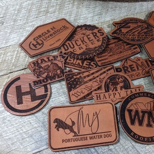 May include: A collection of brown leather patches with various designs and text, including "Circle H Livestock", "Duckers Creek", "Half Mowers", "Bikes", "We We Pea", "Premier", "Happy Tail", "Commercial WM Roofing", "Mavvin", "AL Prestige Construction", "EBikes", "My Portuguese Water Dog", and "Willzy".
