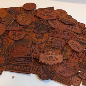 May include: A collection of brown leather patches with various designs and text, including "Dad Daddy", "Duckers Creek", "Holy Bible", "Little Debbie", "Go Broke", and "Rump 2024".