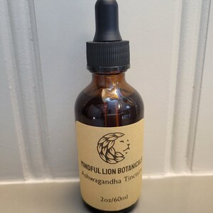Puede incluir: Una botella de vidrio marr&oacute;n con un cuentagotas negro. La botella tiene una etiqueta beige con el texto "Mindful Lion Botanicals Ashwagandha Tincture" y un logotipo de le&oacute;n. Contiene 60 ml de l&iacute;quido.