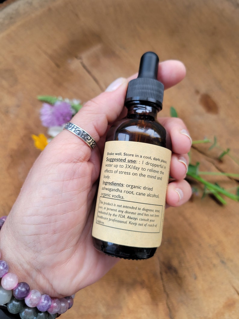 Puede incluir: Una botella de vidrio marr&oacute;n con un cuentagotas negro, sostenida en una mano. La botella tiene una etiqueta con texto sobre ingredientes y uso. El fondo es una superficie de madera con flores.
