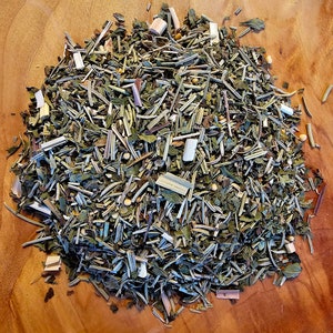 May include: A close-up of a mixture of dried herbs and spices, including green leaves, brown stems, and small white pieces. The mixture is arranged in a pile on a wooden surface.