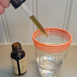 Puede incluir: Un cuentagotas de vidrio transparente dispensa un l&iacute;quido amarillo-marr&oacute;n en un vaso de agua. Una botella de vidrio &aacute;mbar con una etiqueta est&aacute; cerca. El vaso tiene rayas naranjas alrededor del borde. El fondo es una superficie gris claro.