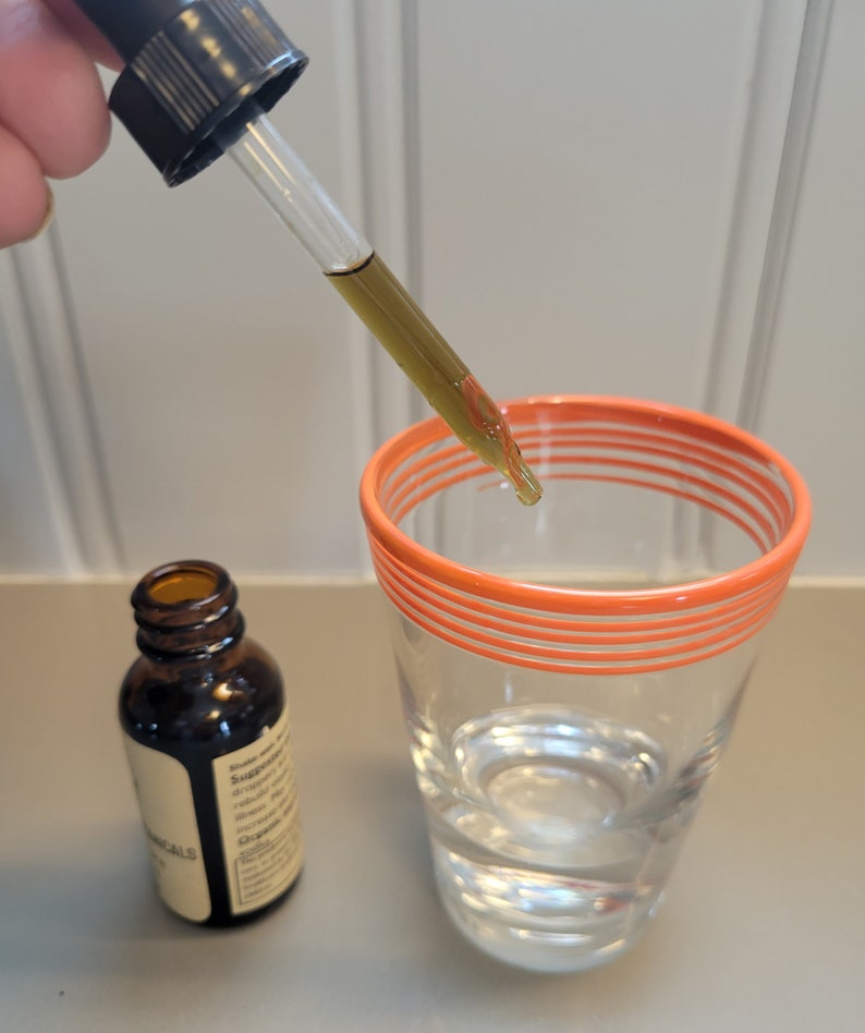 Puede incluir: Un cuentagotas de vidrio transparente dispensa un l&iacute;quido amarillo-marr&oacute;n en un vaso de agua. Una botella de vidrio &aacute;mbar con una etiqueta est&aacute; cerca. El vaso tiene rayas naranjas alrededor del borde. El fondo es una superficie gris claro.
