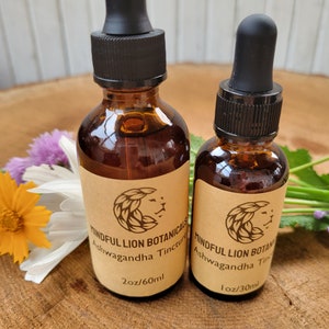 Puede incluir: Dos botellas de vidrio &aacute;mbar de Mindful Lion Botanicals Ashwagandha Tincture con cuentagotas negros. La botella m&aacute;s grande est&aacute; etiquetada 60 ml, y la m&aacute;s peque&ntilde;a es 30 ml. Las etiquetas presentan un logotipo de cabeza de le&oacute;n. Las botellas est&aacute;n sobre una superficie de madera con flores.