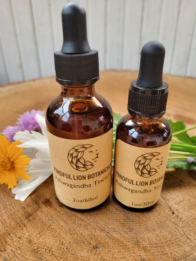Puede incluir: Dos botellas de vidrio &aacute;mbar de Mindful Lion Botanicals Ashwagandha Tincture con cuentagotas negros. La botella m&aacute;s grande est&aacute; etiquetada 60 ml, y la m&aacute;s peque&ntilde;a es 30 ml. Las etiquetas presentan un logotipo de cabeza de le&oacute;n. Las botellas est&aacute;n sobre una superficie de madera con flores.