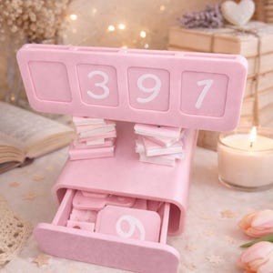 May include: A pink desk organizer with the numbers 397 displayed on top. The organizer has a drawer with the number 6 visible. Small stacks of pink cards are placed on top. A lit candle and flowers are in the background.
