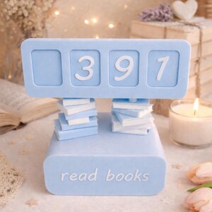 May include: A light blue book counter with the numbers 397 displayed. The counter is supported by two stacks of miniature books and the base reads "read books". The background includes a lit candle, flowers, and fairy lights.