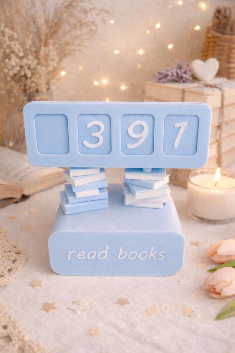 May include: A light blue book counter with the numbers 397 displayed. The counter is supported by two stacks of miniature books and the base reads "read books". The background includes a lit candle, flowers, and fairy lights.