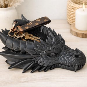 May include: A black dragon head sculpture used as a key holder. A brown and gold patterned keychain and keys are on the dragon's head. The sculpture has detailed scales and sharp features, creating a fantasy theme.