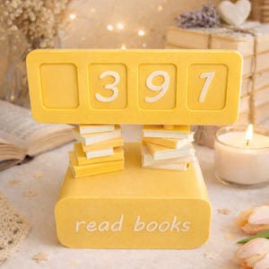May include: A yellow decorative book counter with the numbers 397 displayed. The base reads "read books". Small stacks of book-shaped objects are below the counter. The background includes a lit candle and decorative elements.