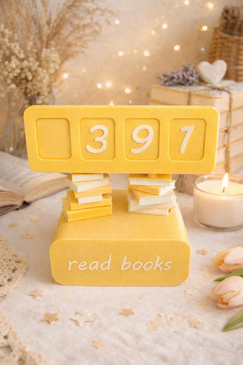 May include: A yellow decorative book counter with the numbers 397 displayed. The base reads "read books". Small stacks of book-shaped objects are below the counter. The background includes a lit candle and decorative elements.
