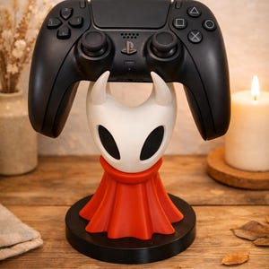 May include: A black games controller sits on a white and red character-themed stand. The stand features a stylized face with horns and a red cape, set on a black circular base. The controller has buttons and joysticks.