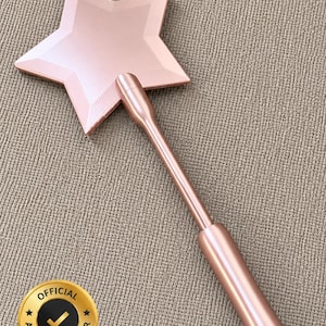 May include: A rose gold star-shaped wand with a long handle. The star has a textured surface and is connected to a cylindrical handle. The wand is on a neutral-colored textured surface. An official seller badge is in the lower left corner.