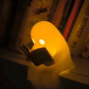 May include: A glowing yellow night light shaped like a ghost reading a book. The light has two eye-shaped cutouts and is positioned on a shelf in front of a row of books. The light emits a warm glow.