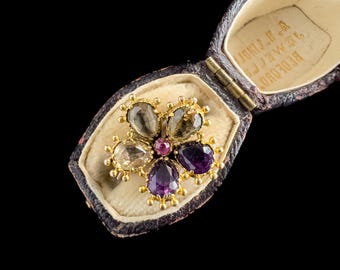 May include: A gold flower brooch with a red center stone and purple and brown stones around it. The brooch is in a small, brown velvet box with the inscription "REDFERN & JAMES LTD."