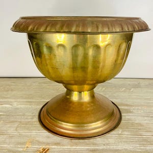 May include: A vintage brass planter with a wide, fluted bowl and a pedestal base. The planter has a warm, golden hue with a slightly aged appearance. The rim has a darker, bronze-like color. The planter is designed to hold plants and flowers.