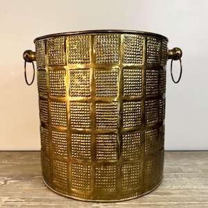 May include: A cylindrical brass bucket featuring a grid pattern of raised, textured squares across its exterior surface. The vessel includes two side-mounted metal rings attached to small knobs, providing handles for lifting the container.
