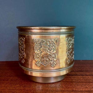 Peut inclure: Un cache-pot cylindrique en laiton avec une finition métallique polie et un motif en relief répété. Le design présente un blason héraldique central avec un casque et une fleur de lys, entouré de feuillages sculptés complexes.