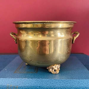 May include: A vintage brass pot with two small handles and three decorative feet. The pot has a rounded body and a wide rim. The brass has a warm, golden colour and shows signs of age. The pot is sitting on a blue surface against a red background.