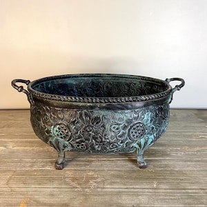 May include: An antique bronze-colored planter with a detailed floral pattern. The oval-shaped bowl has two handles and rests on three small feet. The planter has a weathered, aged appearance with hints of blue-green patina.