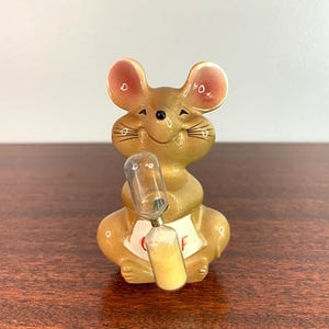 May include: A glossy tan ceramic mouse figurine with large ears and a smiling expression, holding a clear glass hourglass filled with light yellow sand. The mouse features a white apron on its front with the red text "CHEF" printed on it.