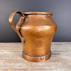 May include: A copper pitcher with a wide, rounded body and a single curved handle attached to the side. The metal surface features a hammered texture with visible seams and rivets securing the handle to the main body of the vessel.