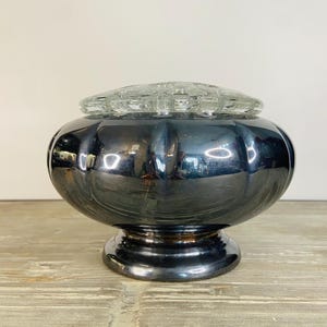 May include: A vintage, silver-toned vase with a round, ribbed body and a clear, glass flower frog lid. The vase sits on a pedestal base. The reflective surface of the vase shows the surrounding environment. The vase is likely made of metal.