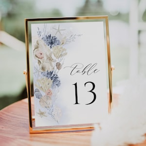 May include: A framed table number card with a gold-colored frame. The card features a watercolor design of seashells, starfish, and coral in shades of blue, beige, and peach. The number "13" is displayed in a bold black font.